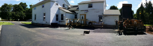 Workers pave a driveway with asphalt in front of a white house on a sunny day.