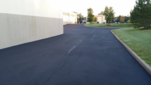 Freshly paved asphalt driveway curves past a beige building with trees and grass nearby.