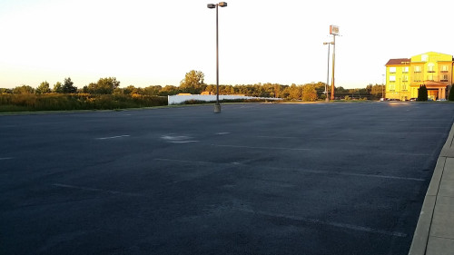 Empty parking lot with a few light poles, a distant building, and trees in the background.