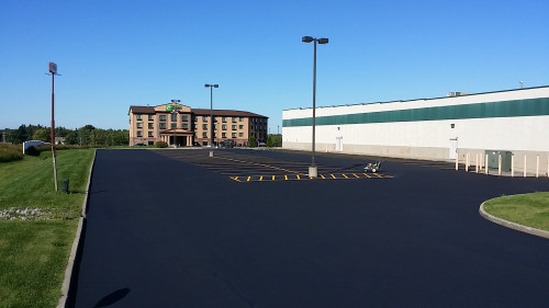Freshly paved empty parking lot with a few lamp posts, next to a hotel and a large white building.