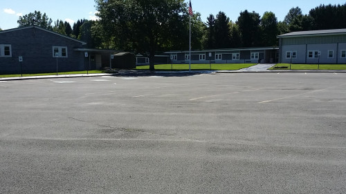 Empty parking lot in front of a single-story school building with trees and flagpole in view.