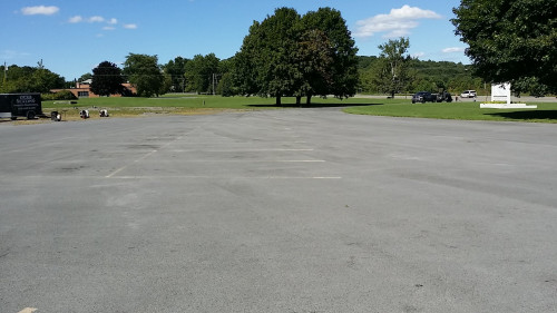 Empty parking lot with trees and a grassy field in the background under a blue sky.