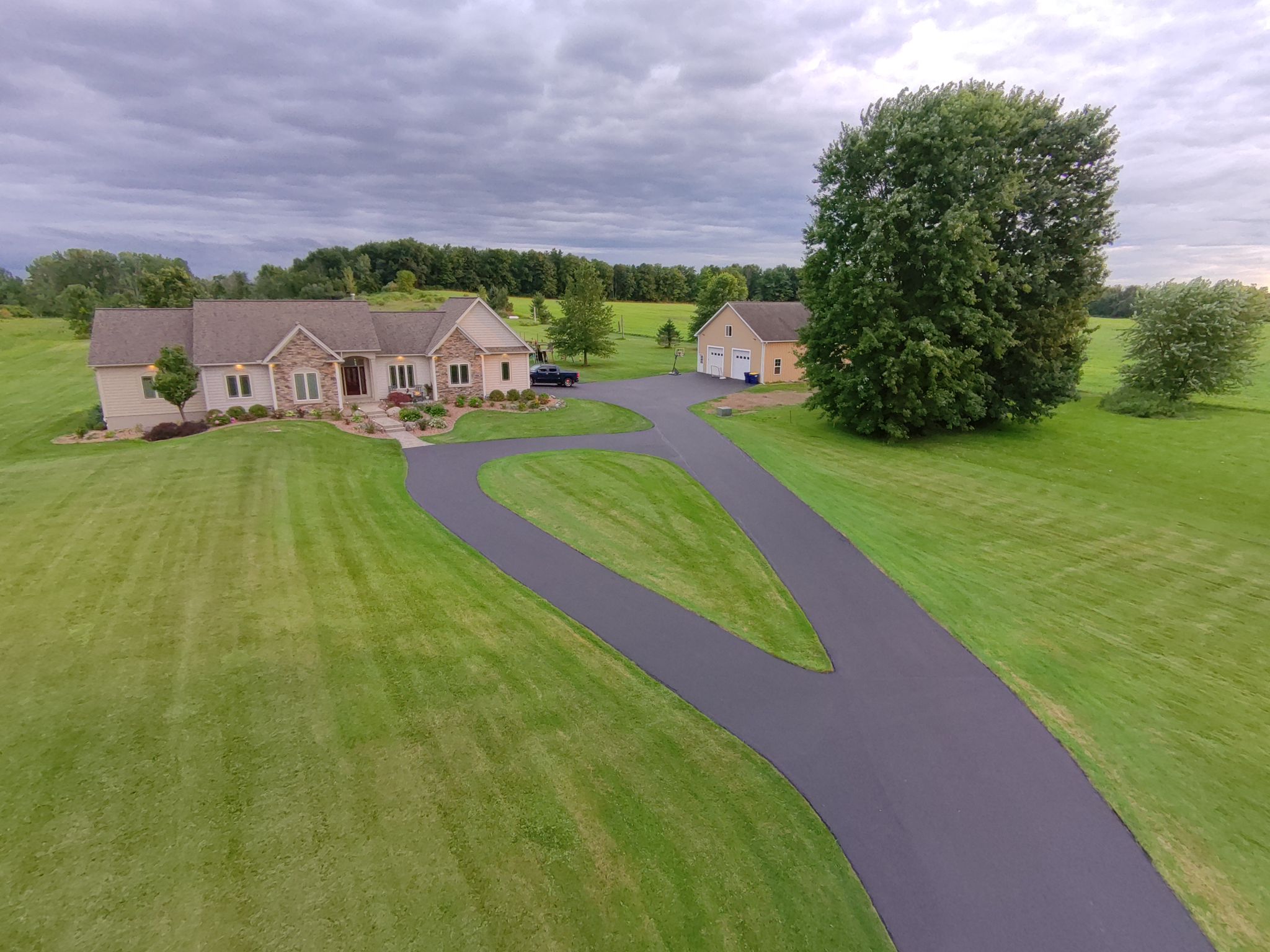 A house with a detached garage, long driveway, and large green lawn under a cloudy sky.