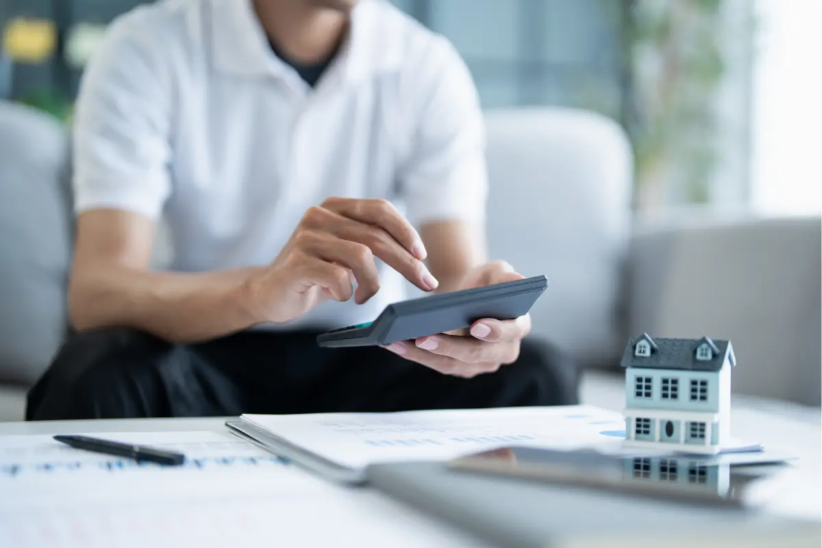 A person uses a calculator near documents and a small model house on a table.