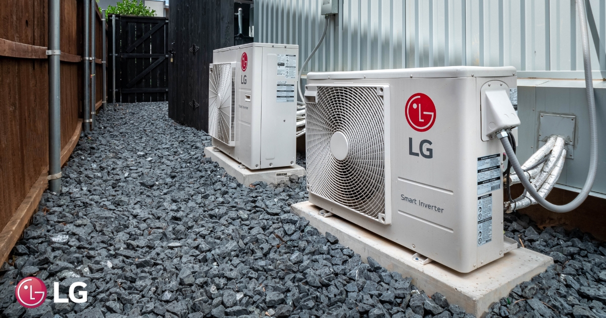 Two LG outdoor air conditioning units sit on gravel beside a building and wooden fence.