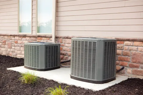 Two outdoor air conditioning units sit on a concrete pad next to a house with siding and brick.