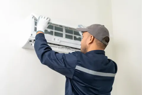 A technician in uniform servicing a wall-mounted air conditioner unit indoors.