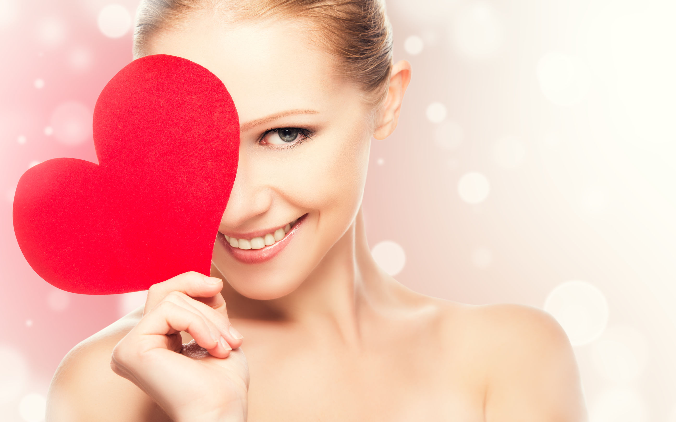 Smiling woman holding a red paper heart over one eye, with a soft pink blurred background.