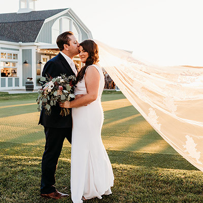 couple stands on grass, with the groom kissing the brides forehead, her veil flowing in the wind