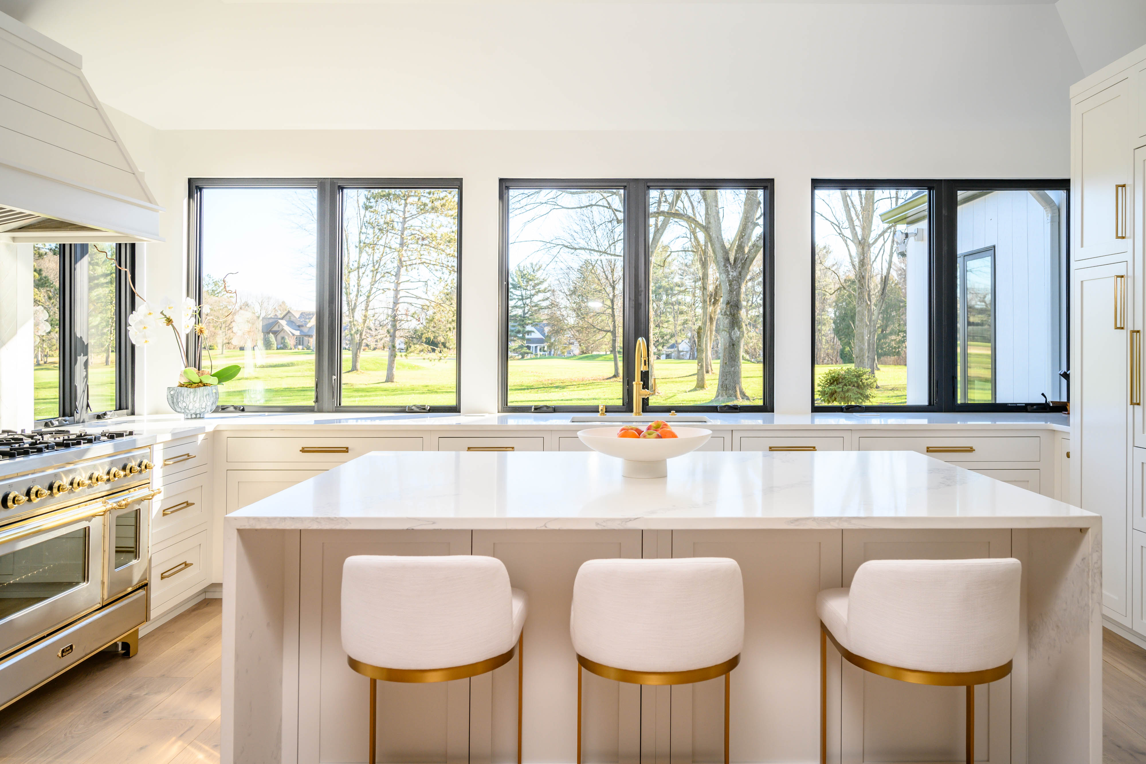 Bright modern kitchen with white island, gold accents, and large windows overlooking a green lawn.