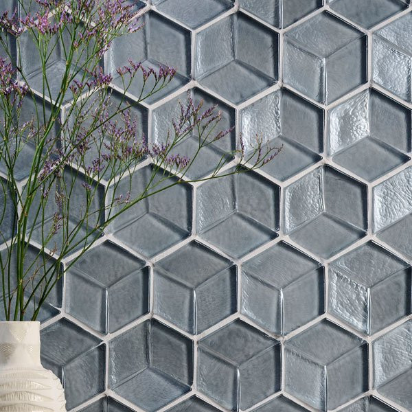 A white vase with purple flowers in front of a blue-gray hexagon tile wall.