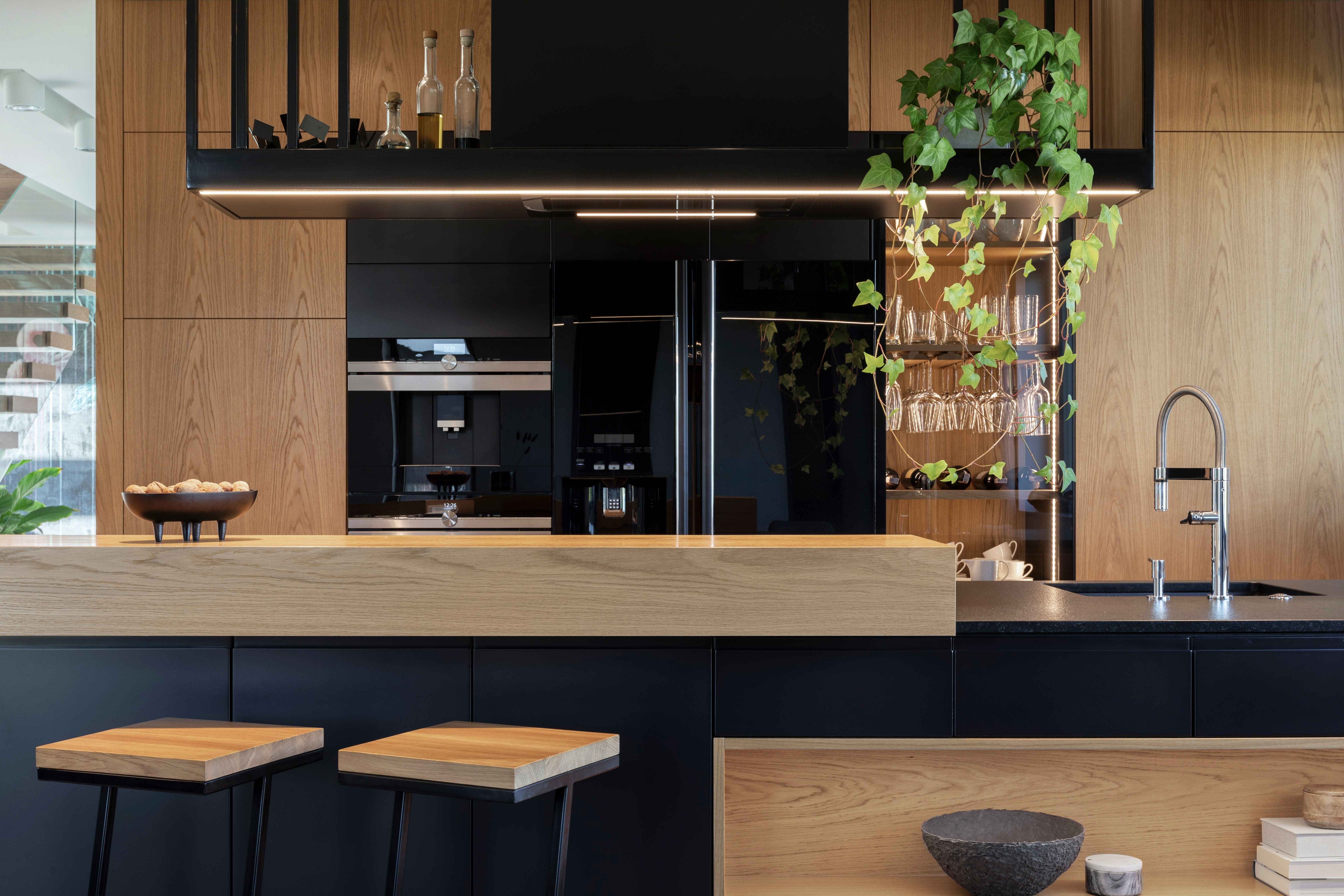 Modern kitchen with wood and black finishes, island seating, plants, and built-in appliances.