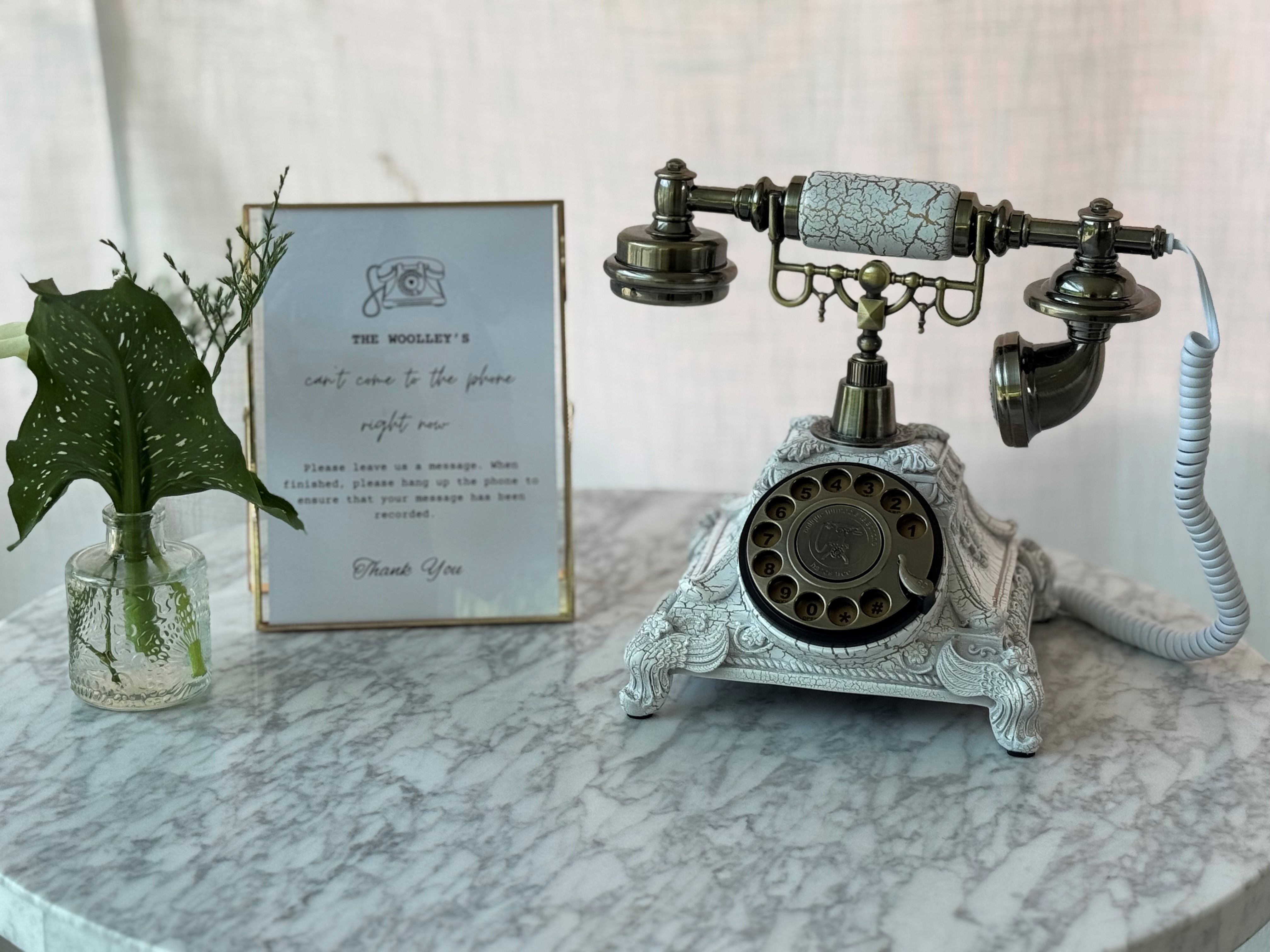 Vintage rotary telephone and a framed note sit on a marble table next to a small vase of leaves.