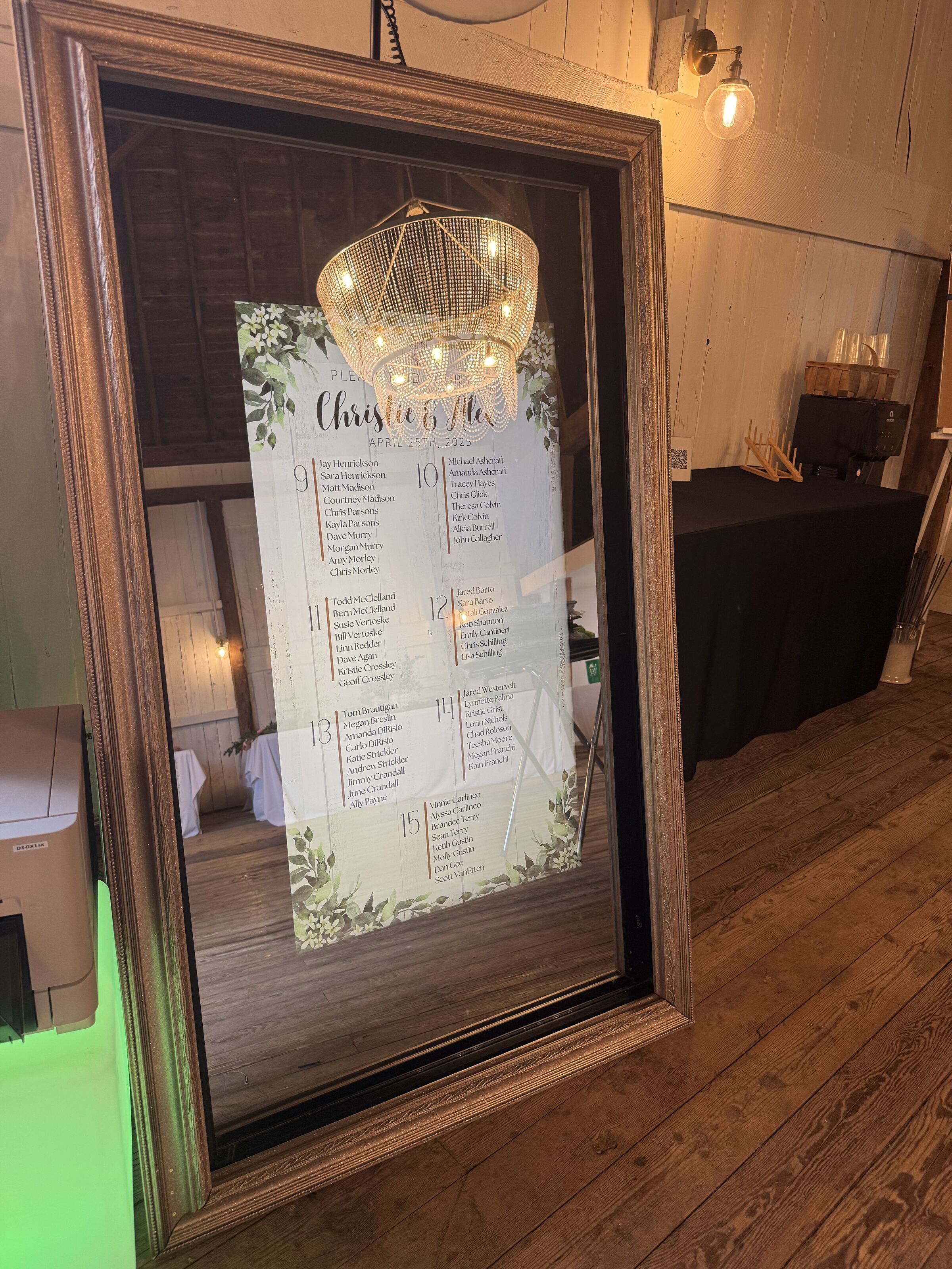 A mirror photo booth displays a wedding seating chart against a wall in a rustic wooden venue.