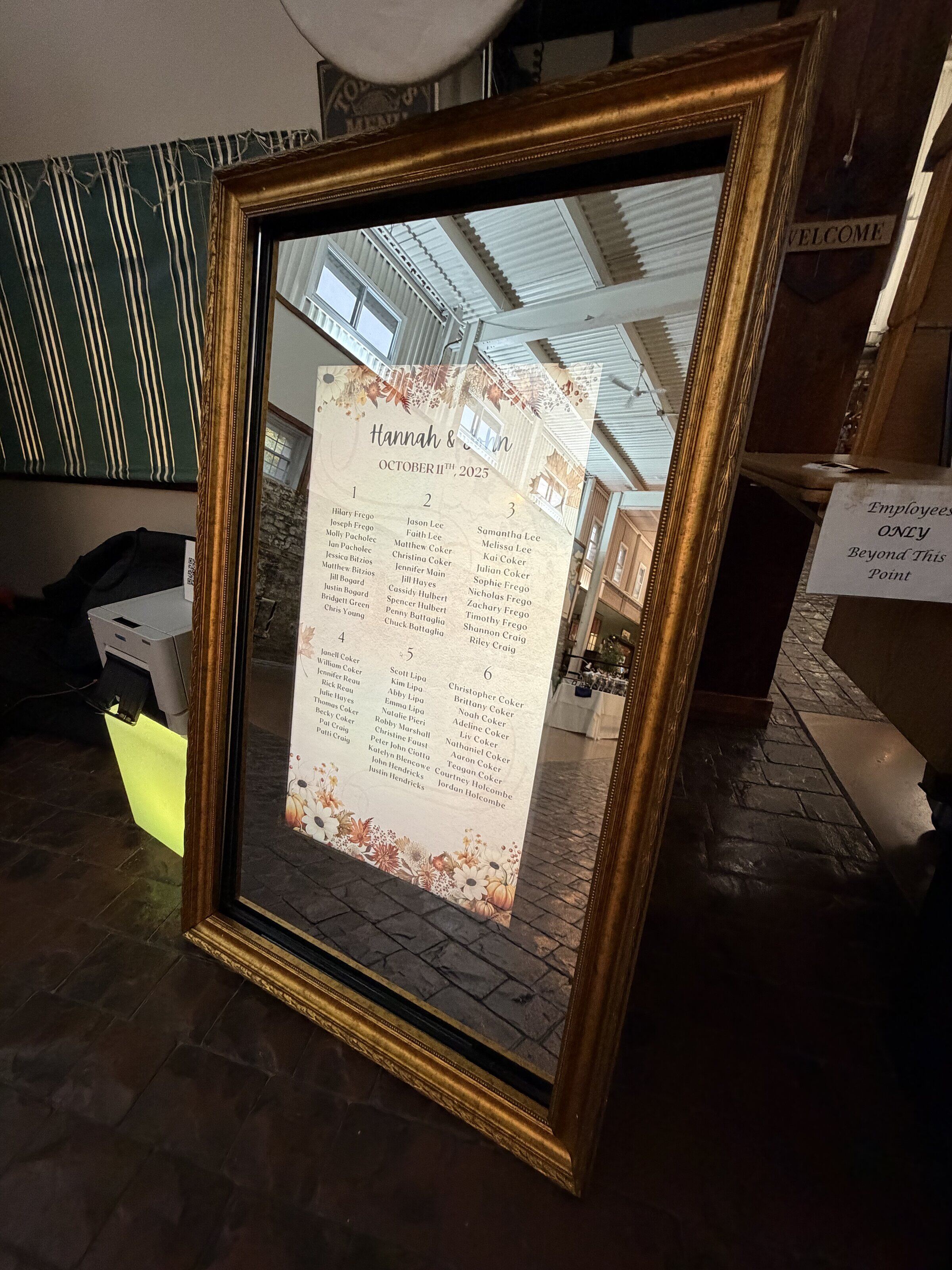 A large framed wedding seating chart stands on a wooden floor, reflecting the room around it.
