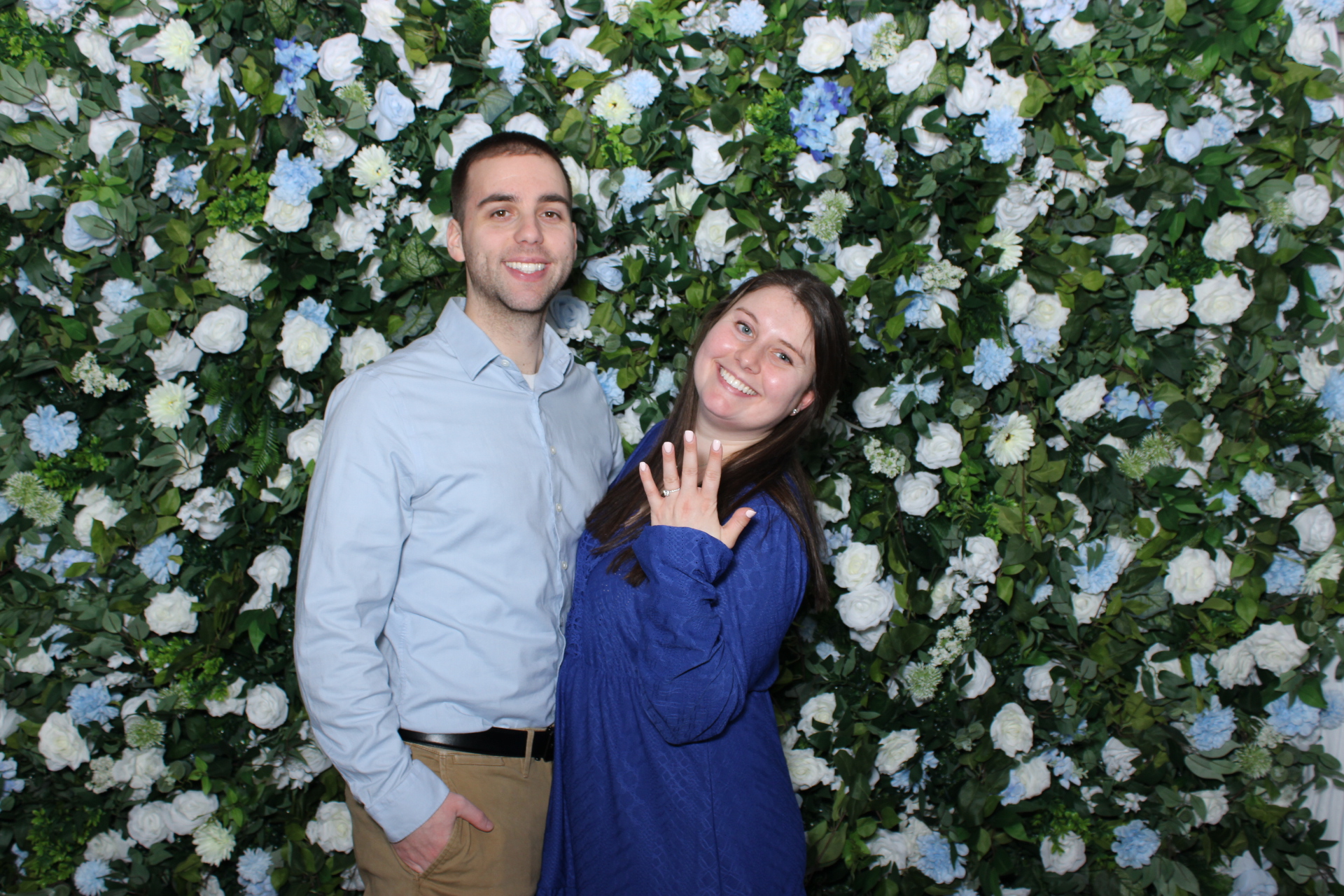 Couple in front of Something Blue flower wall backdrop at engagement celebration