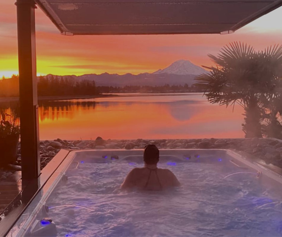 woman in swim spa overlooking a lake with Mt. Hood and a pink sunset.
