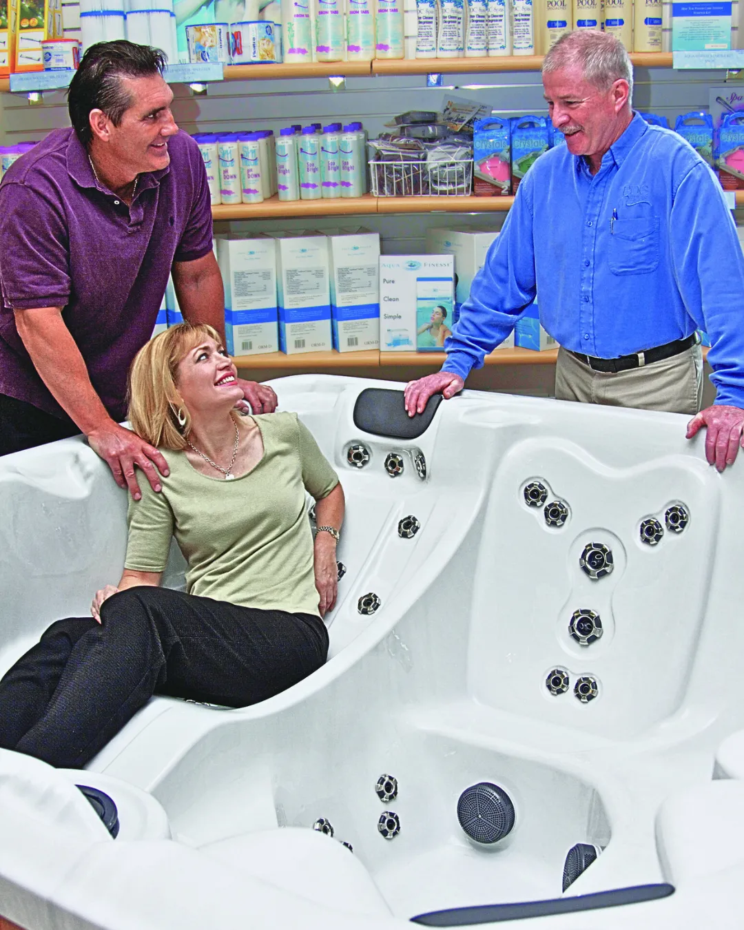 pdc spas showroom with a man leaning over wife who is sitting in an empty hot tub seat. PDC Spas staff memmber speaking with them.
