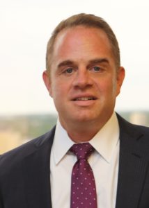 A man in a suit and purple polka dot tie, smiling in front of a blurred background.