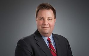 Man in a dark suit, red striped tie, and light blue shirt, smiling against a gray background.