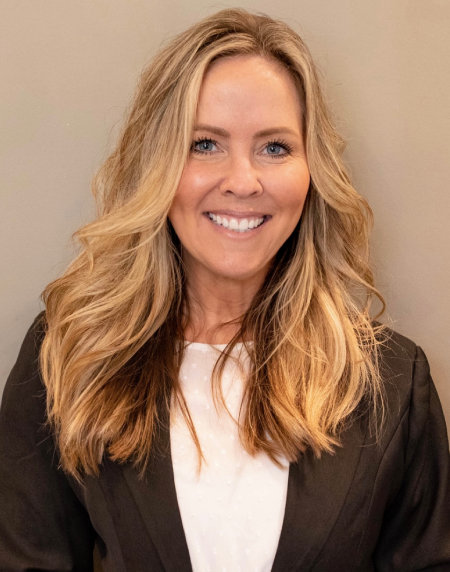Smiling woman with long blonde hair wearing a black blazer and white top, against a tan background.