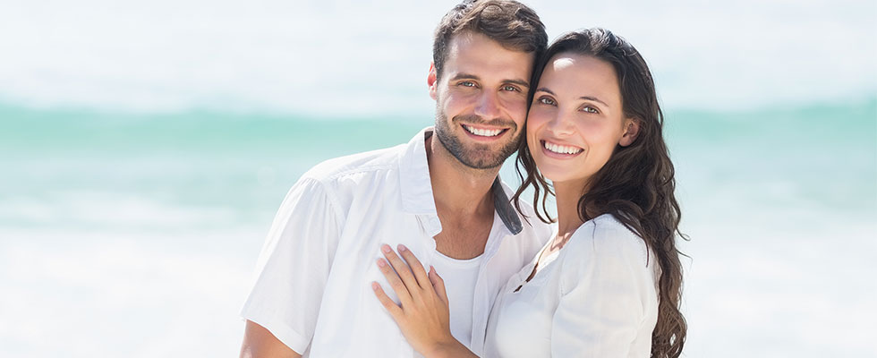 Smiling couple standing close together on a beach with the ocean in the background.
