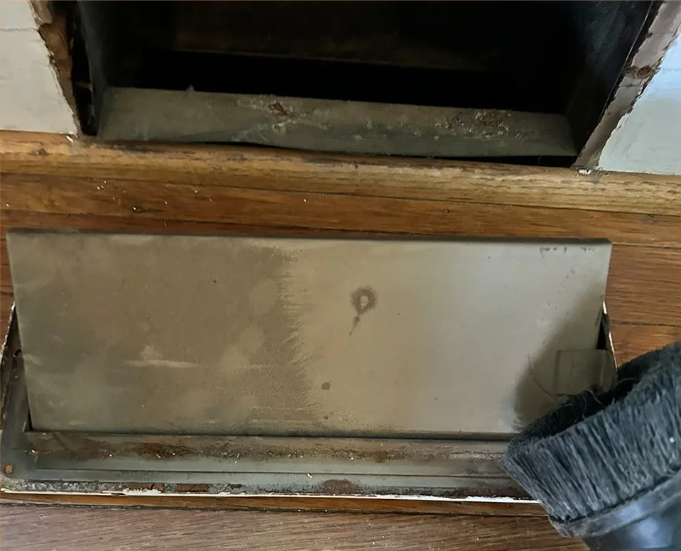 A dirty air vent on a wooden floor with a brush on the righthand side.