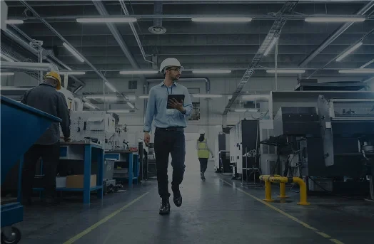 Man in hard hat walks through factory holding tablet; other workers and machinery are visible.