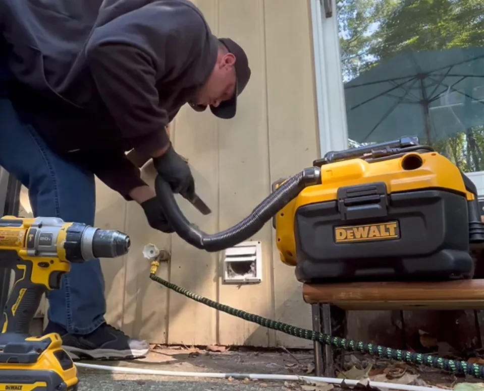 A man leaning over using a DEWALT machine to clean a dryer vent outside of a house.