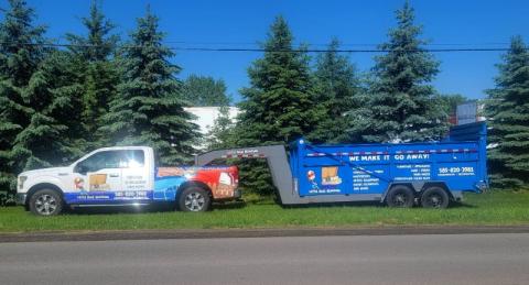 A white truck towing a blue junk removal trailer parked on grass by pine trees.