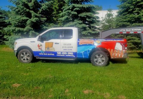 White and blue pickup truck with moving company branding parked on green grass near trees.