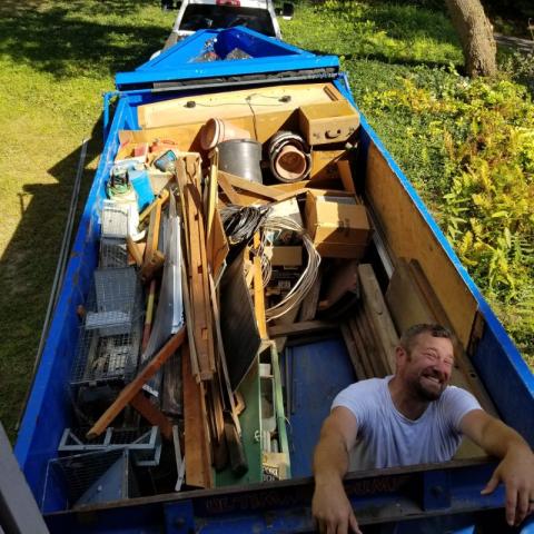 Man smiles while standing in a large dumpster filled with various scrap and household items.