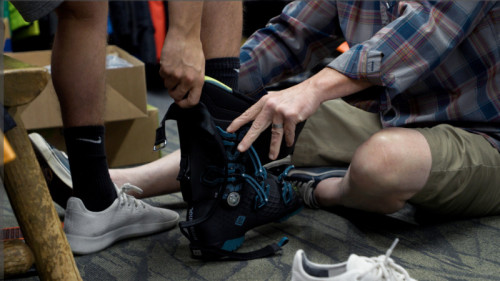 man in plaid shirt holds ski boot while man puts his foot in it