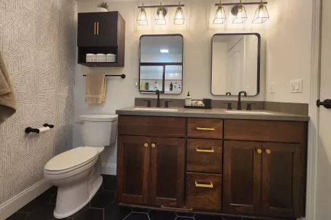 Modern bathroom with double sinks, wood cabinets, wall shelves, and black hexagonal floor tiles.