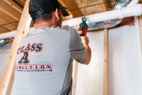 Man in a Class A Construction shirt uses a power drill on a white pipe in a basement.
