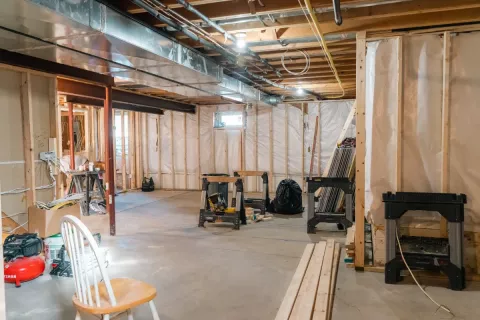 Unfinished basement with exposed beams, tools, plastic-covered walls, and scattered equipment.
