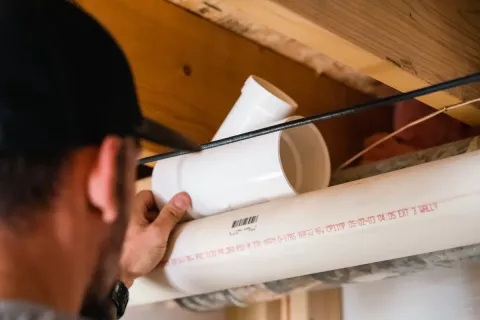 Person installing a white PVC pipe fitting under a wooden ceiling with visible wiring nearby.