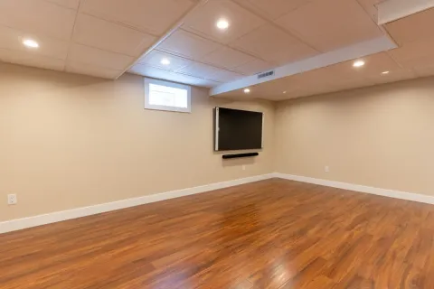 Spacious finished basement with wood flooring, beige walls, recessed lights, and a wall-mounted TV.