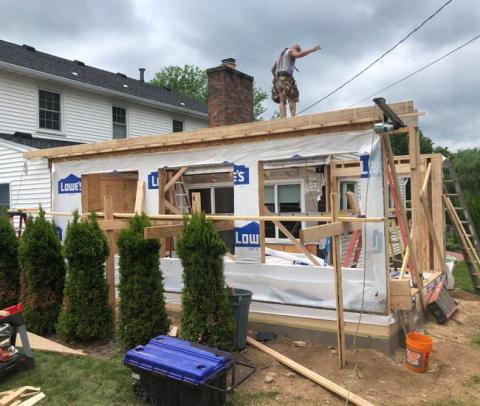 A person works on the roof of a house under construction near some small evergreen trees.