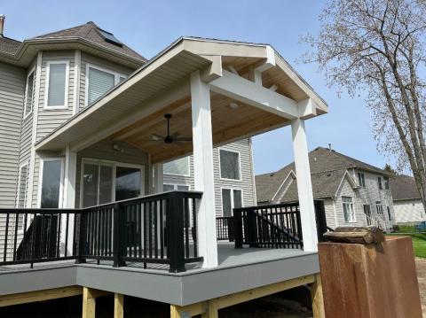 A covered backyard deck with black railings attached to a light gray house on a sunny day.