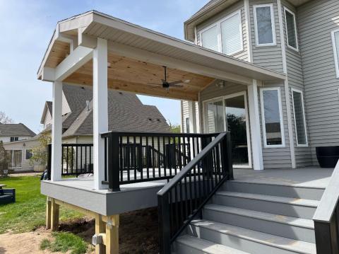 Covered backyard deck with stairs, black railings, and ceiling fan attached to a gray house.