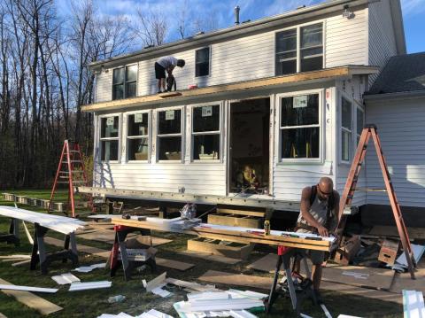 Two people work on the exterior of a white house under renovation, tools and materials scattered.