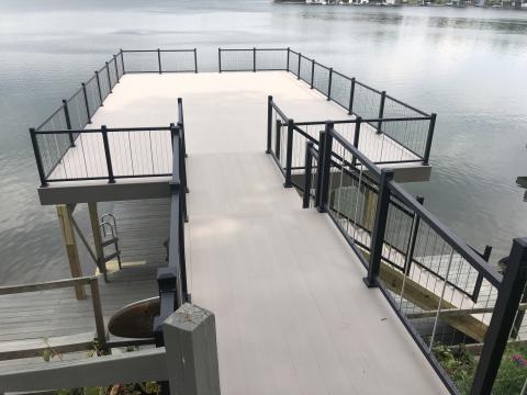 Wooden dock with black railings extending over calm water, seen from a ramp above.