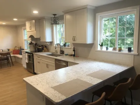 Bright kitchen with white cabinets, marble counters, and bar stools by a large window.