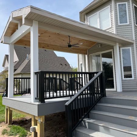 Covered porch with black railing, gray steps, and a ceiling fan attached to a gray house.