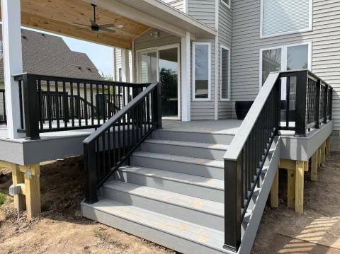 Gray deck with black railings, wide stairs, and a covered area attached to a modern house.