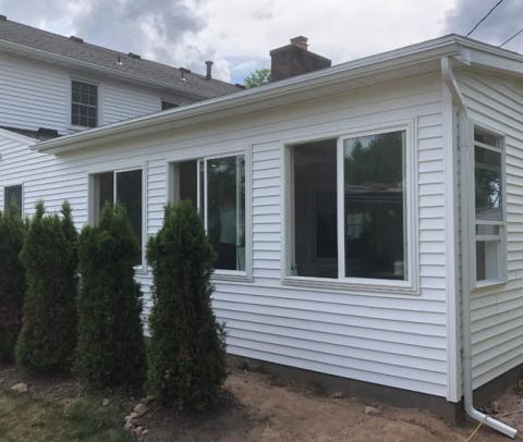 White house with large windows and a sunroom, next to several tall evergreen shrubs.
