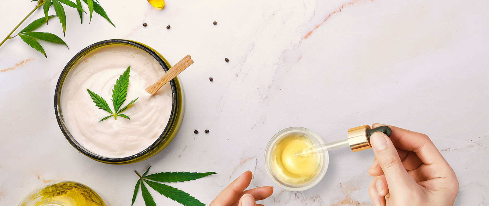 Hands hold a dropper near a cream bowl with a cannabis leaf on a white marble surface.