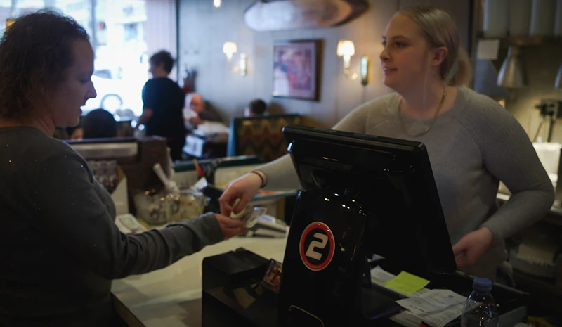 A cashier hands a card back to a customer at a checkout counter in a busy cafe.