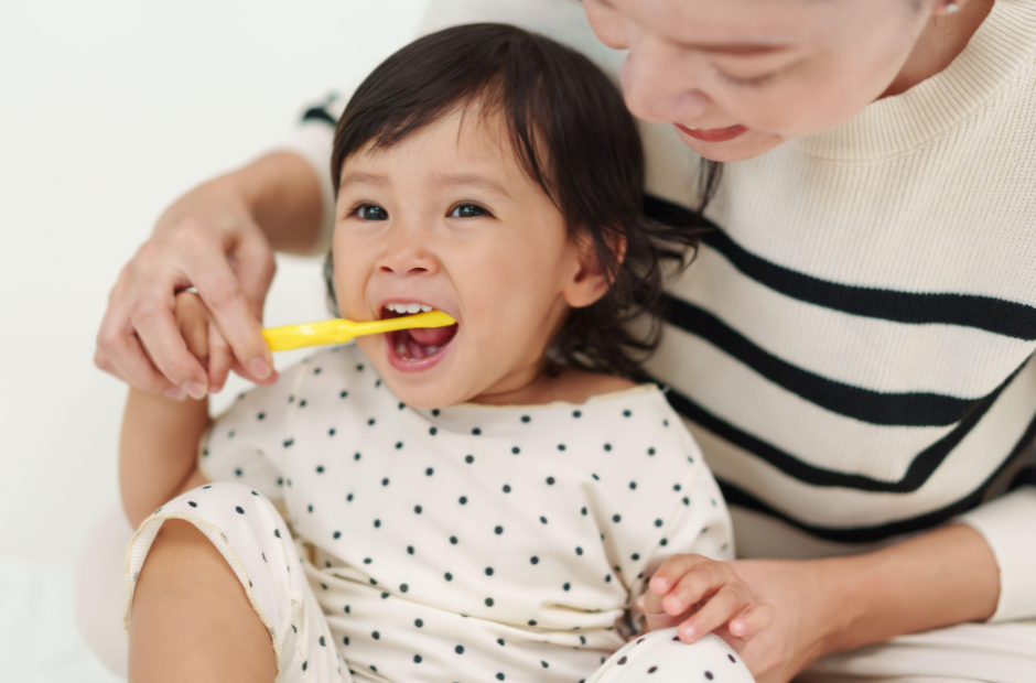 A toddler brushes their teeth with help from an adult, both wearing polka dot and striped clothes.